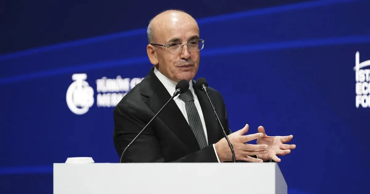 Kürsüde mikrofonlar önünde konuşma yapan gözlüklü ve takım elbiseli Mehmet Şimşek